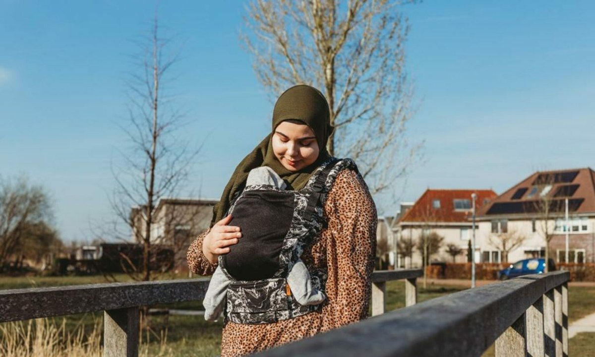 Feride staat bij een houten hek en draagt haar baby in een draagzak op de buik