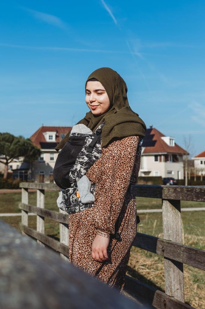 Feride staat bij een houten hek en draagt haar kind in een draagzak op de buik