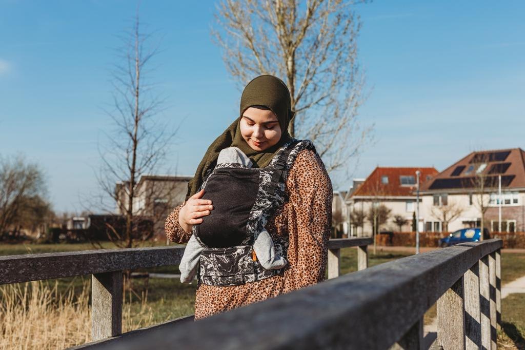 Feride bij een houten hek in de zon met haar baby in een draagzak op de buik