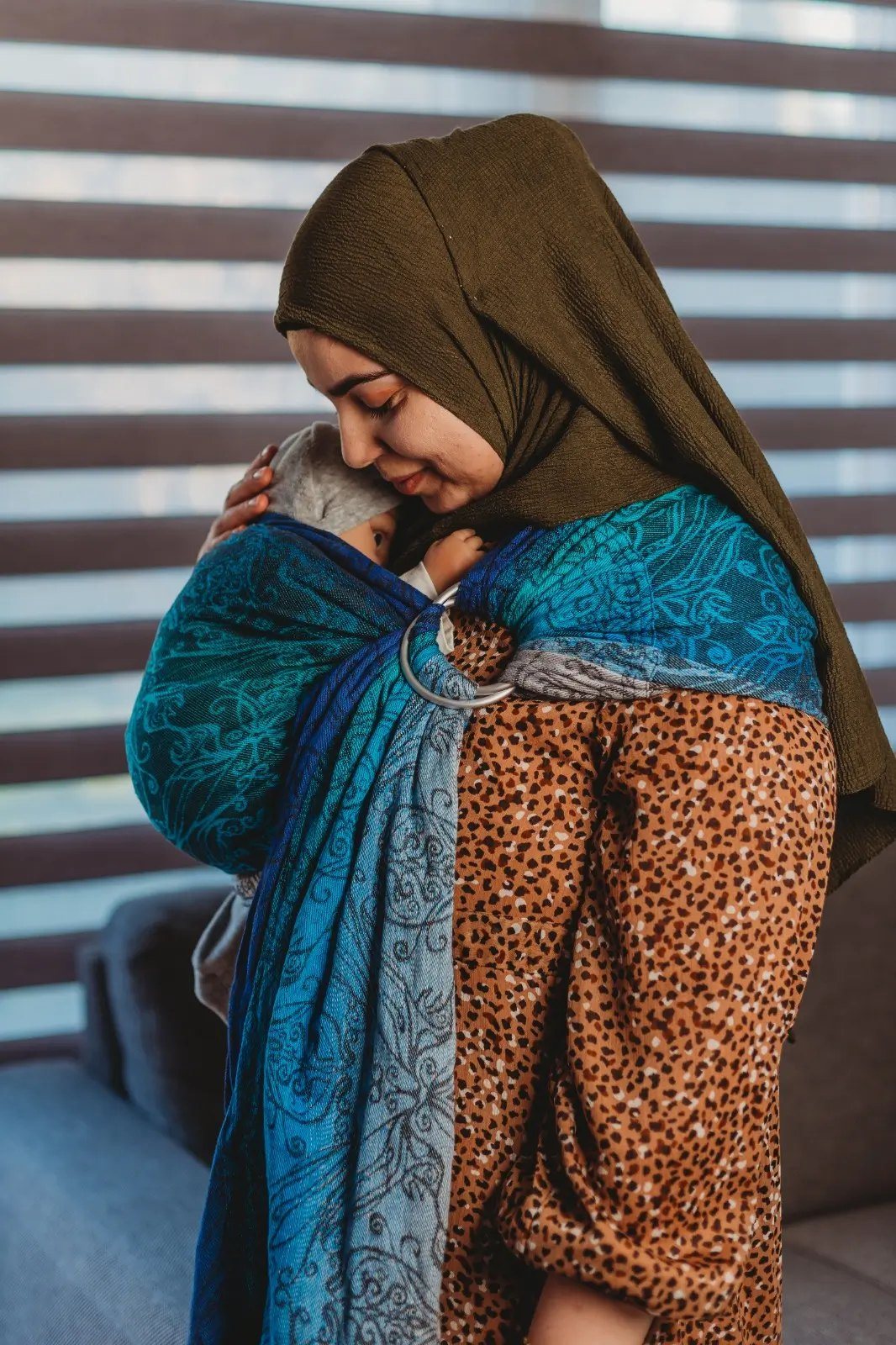Feride houdt haar baby dicht tegen zich aan in een blauwe geweven ringsling, binnenshuis
